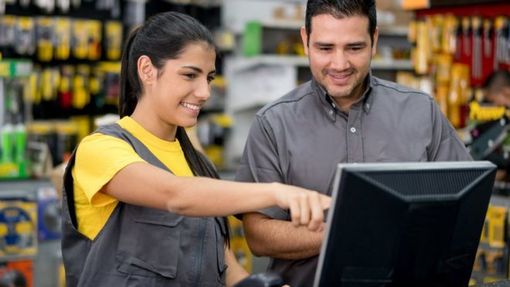 Una donna in camicia gialla e tuta indica lo schermo di un computer insieme a un uomo in camicia grigia in un negozio di ferramenta. Entrambi sorridono.