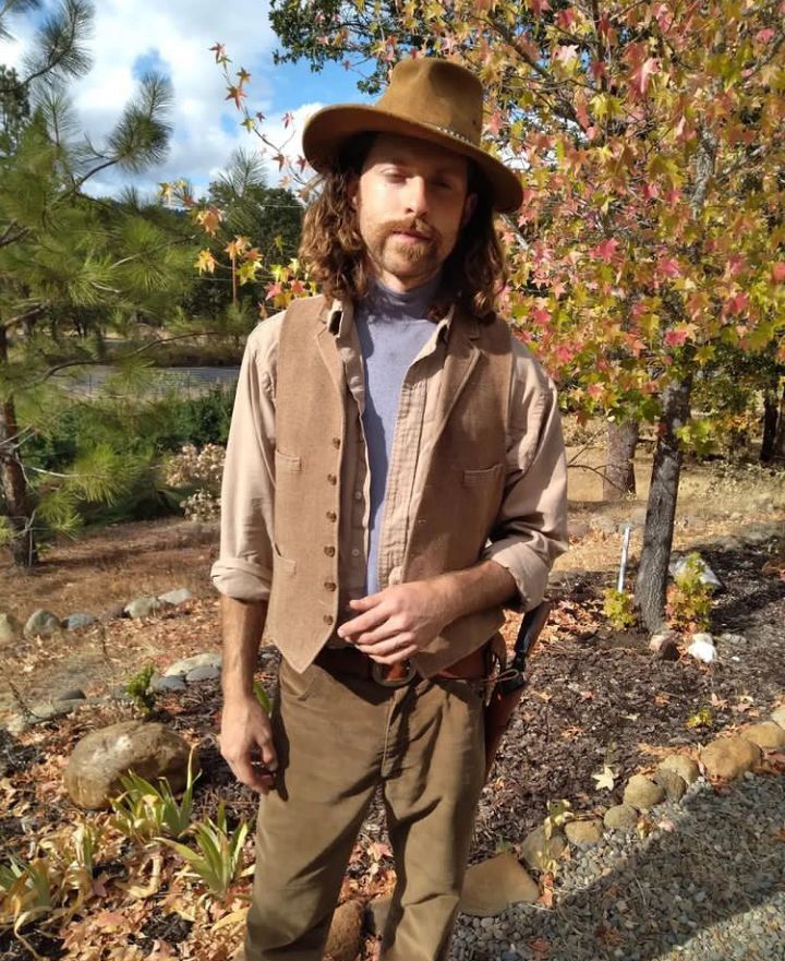 A man in a cowboy hat and vest stands outdoors, posing in front of autumn foliage. He wears a gun holster.