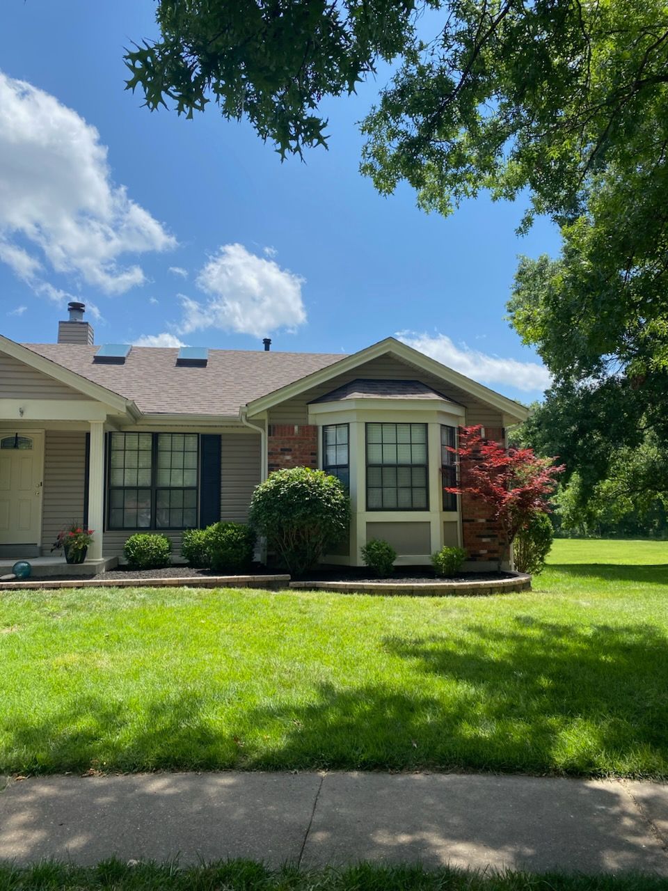 A single-story house with a bay window is surrounded by green grass and trees under a blue sky.