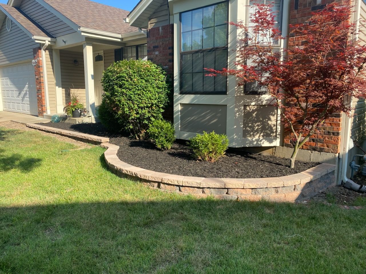 A well-maintained suburban house with a small brick retaining wall and landscaped flower beds with black mulch.
