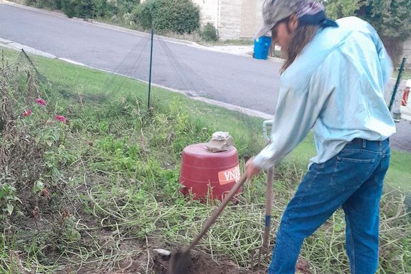 A person with long hair and a camo hat digs in the soil outdoors, near a road and a red container.