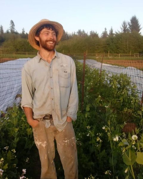 A smiling farmer wearing a straw hat and work clothes stands in a garden, hands in pockets. Green plants fill the foreground.