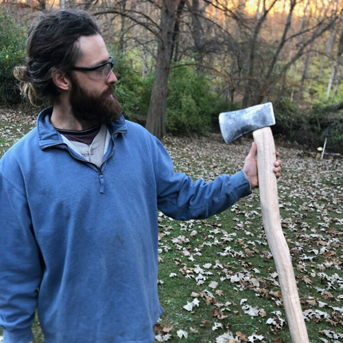 Man with beard and glasses holding a large ax outdoors. 