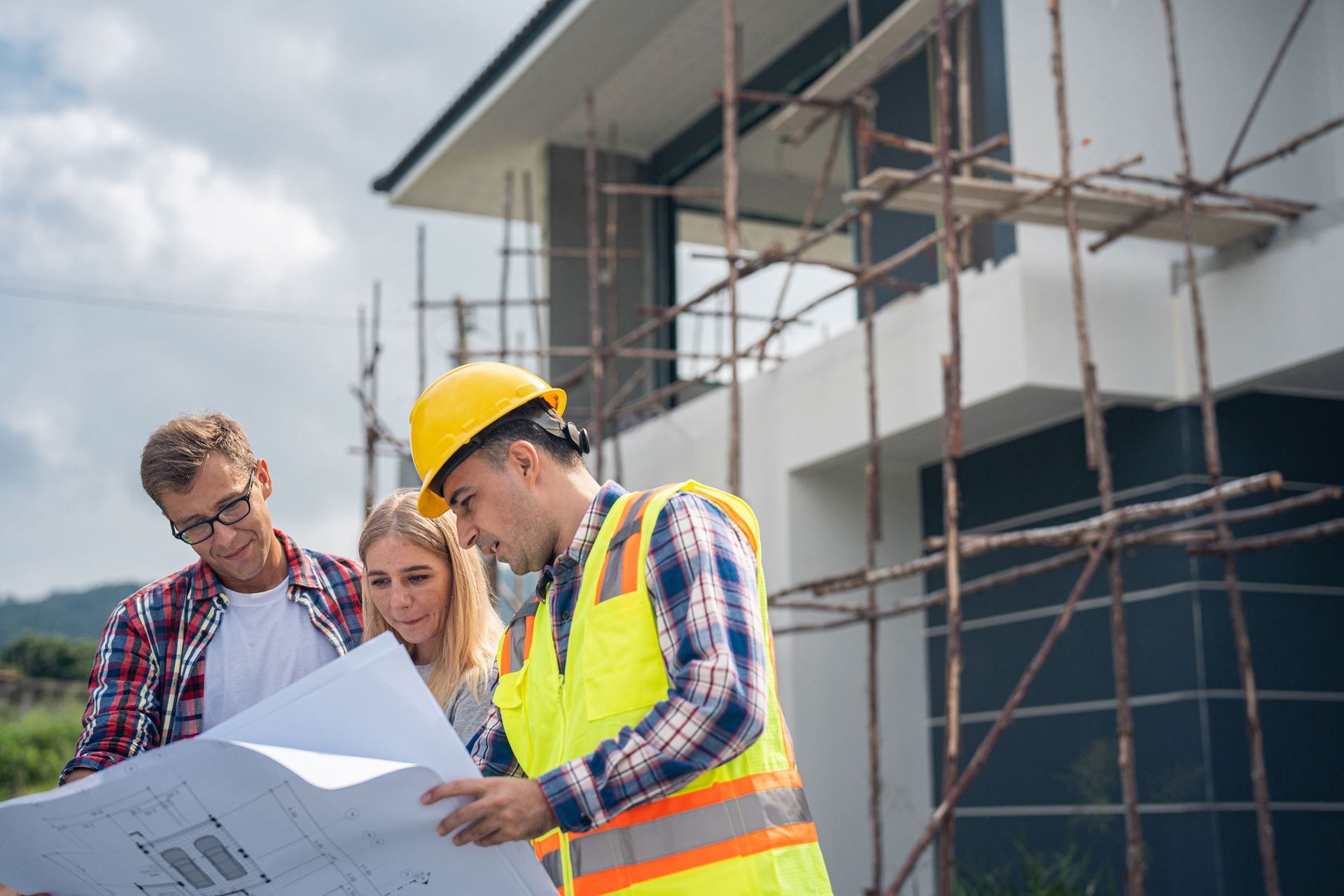 A construction worker in safety gear reviewing blueprints with a couple in front of a building site