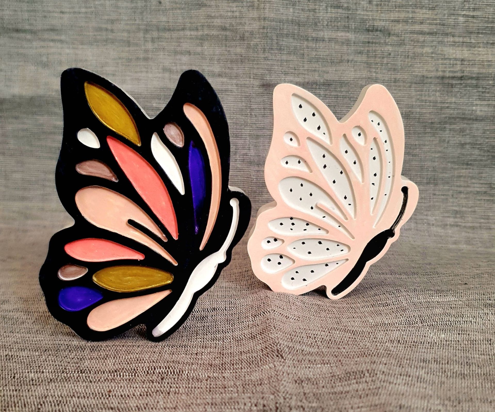 Deux broches décoratives en forme de papillon, l'une noire à émail coloré et l'autre rose pâle, sur une surface en bois
