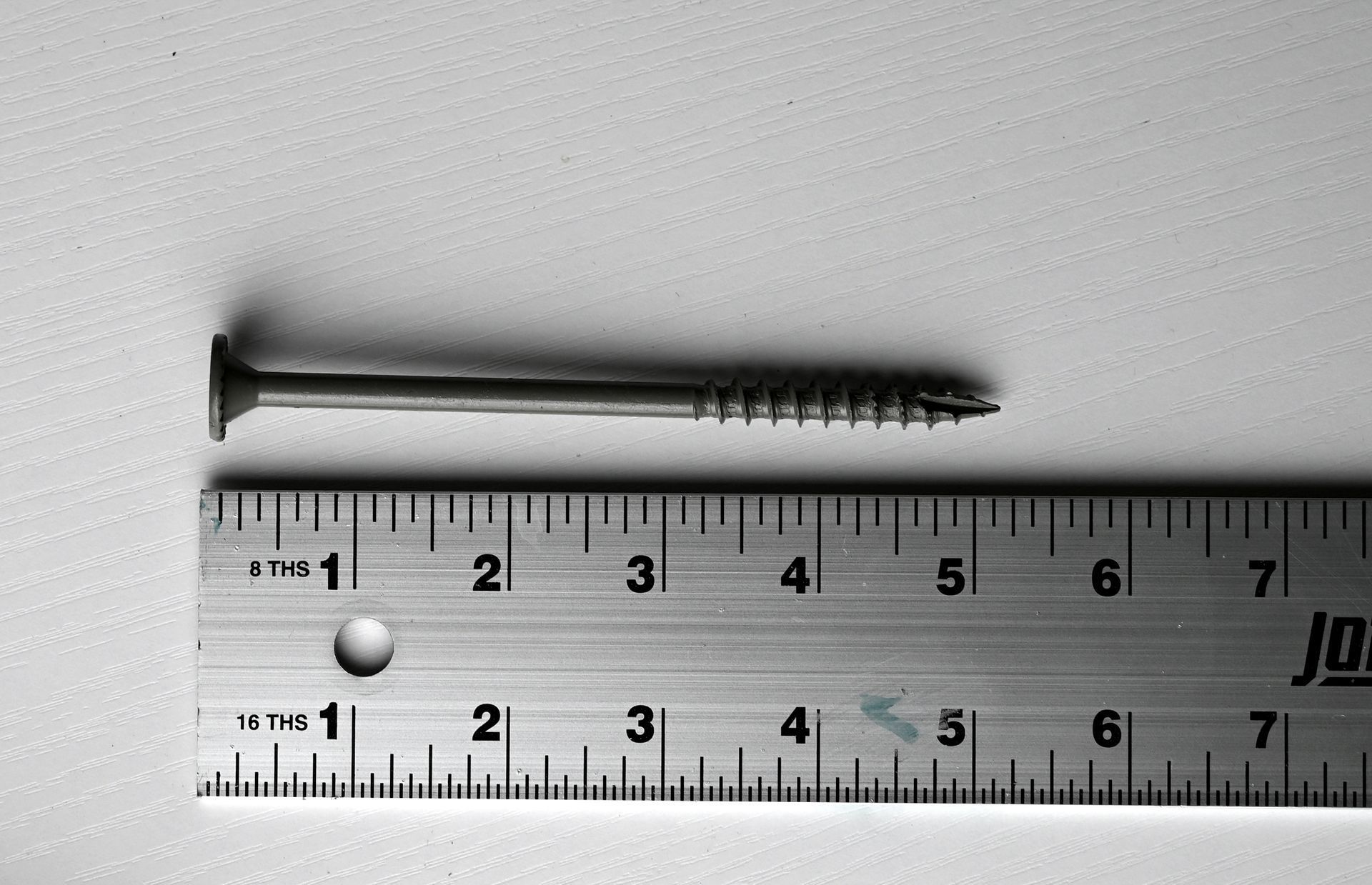 Black screw above a ruler on a white surface, showing its length.