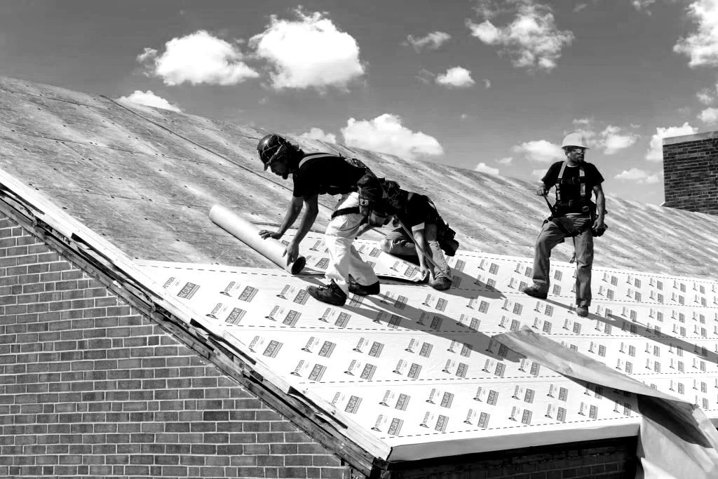 Two workers on a roof installing roofing underlayment under a partly cloudy sky