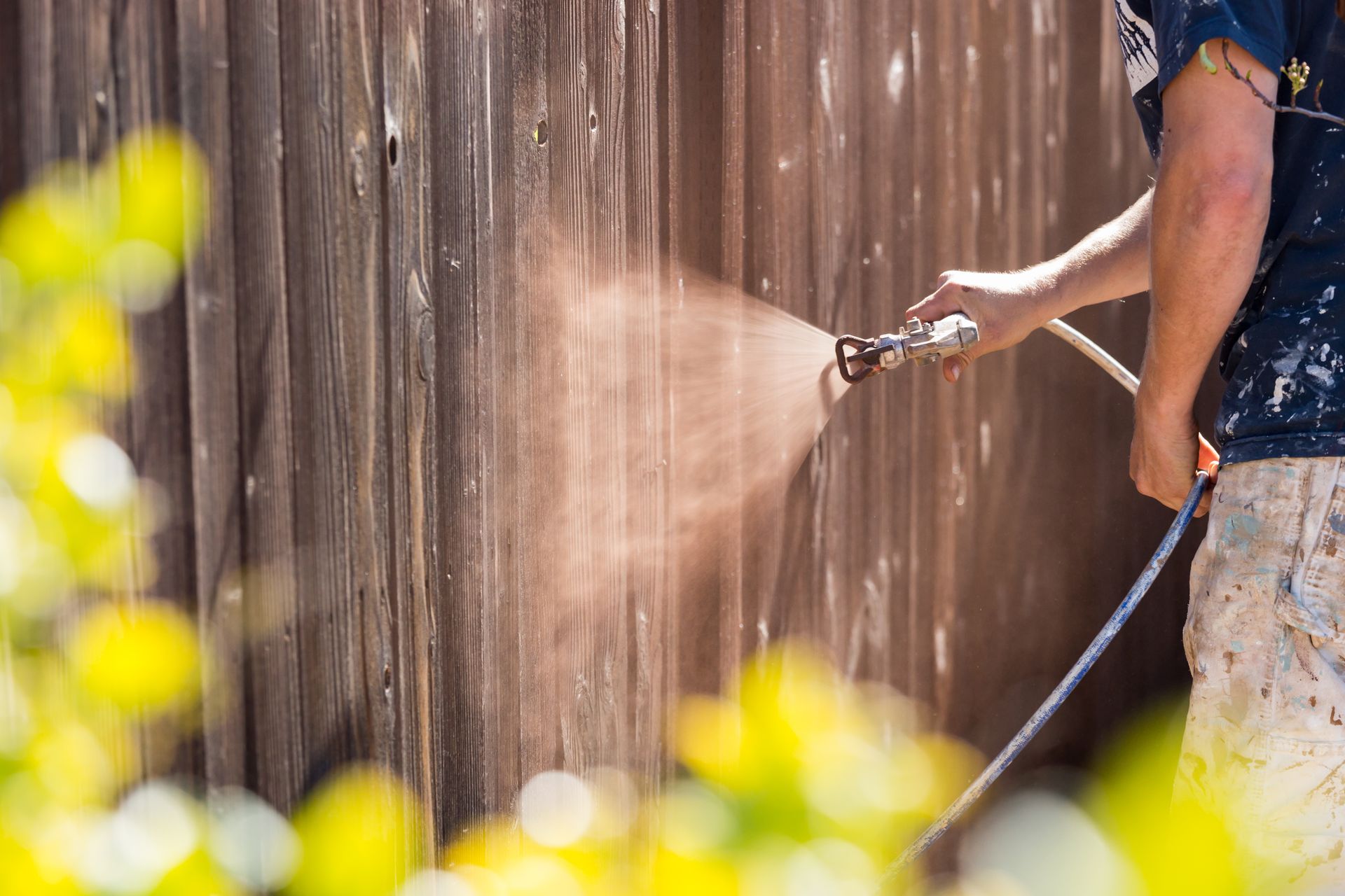 A person using an air spray gun to paint a fence, creating a fine mist of paint particles in the air.