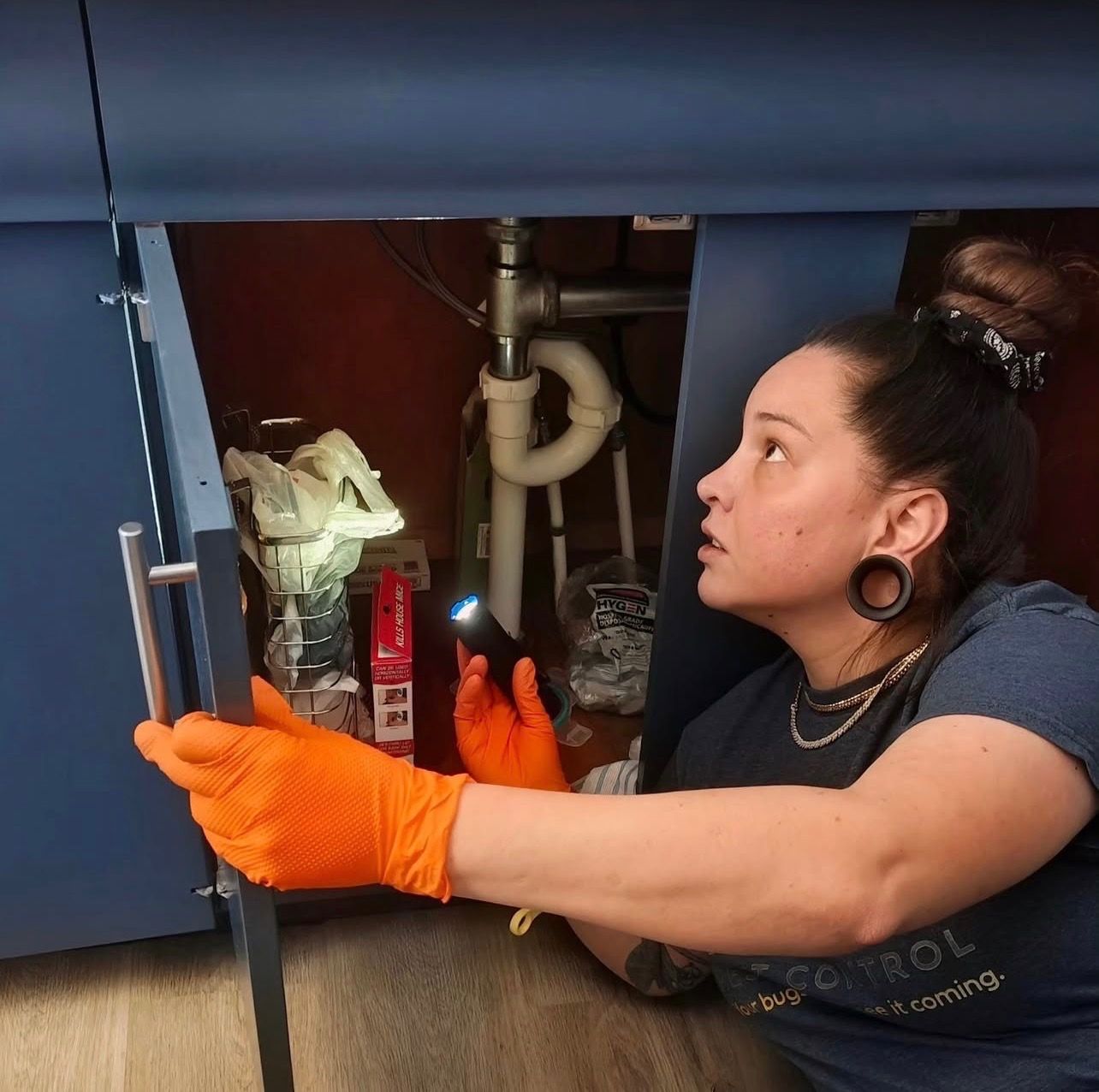 Woman in orange gloves inspecting pipes under a sink with a flashlight