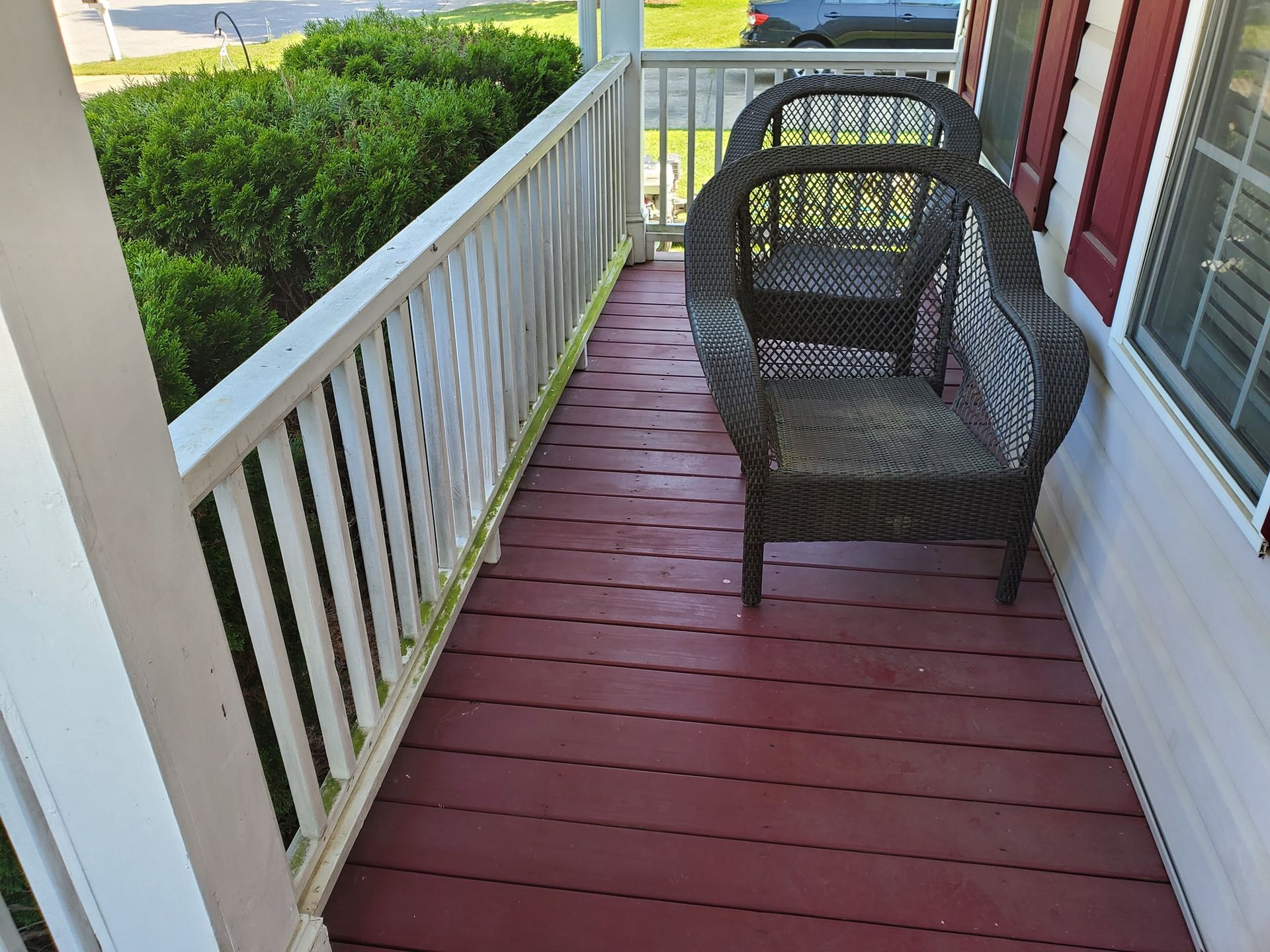 A small porch with two wicker chairs on red wooden planks. White railing and a green bush are visible.