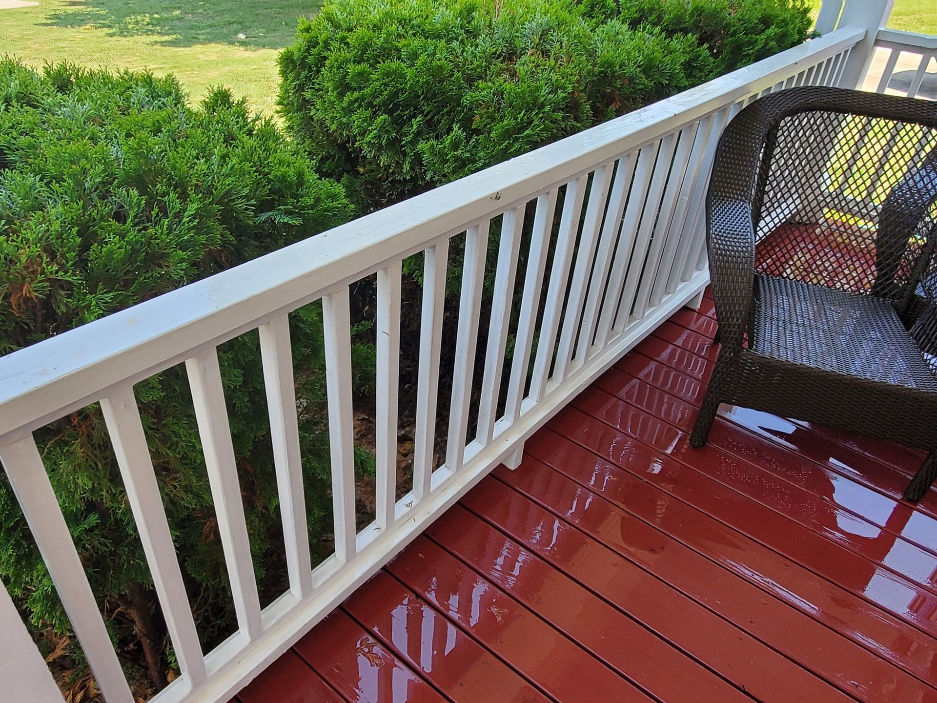 White railing and red deck next to green bushes and a wicker chair on a porch.