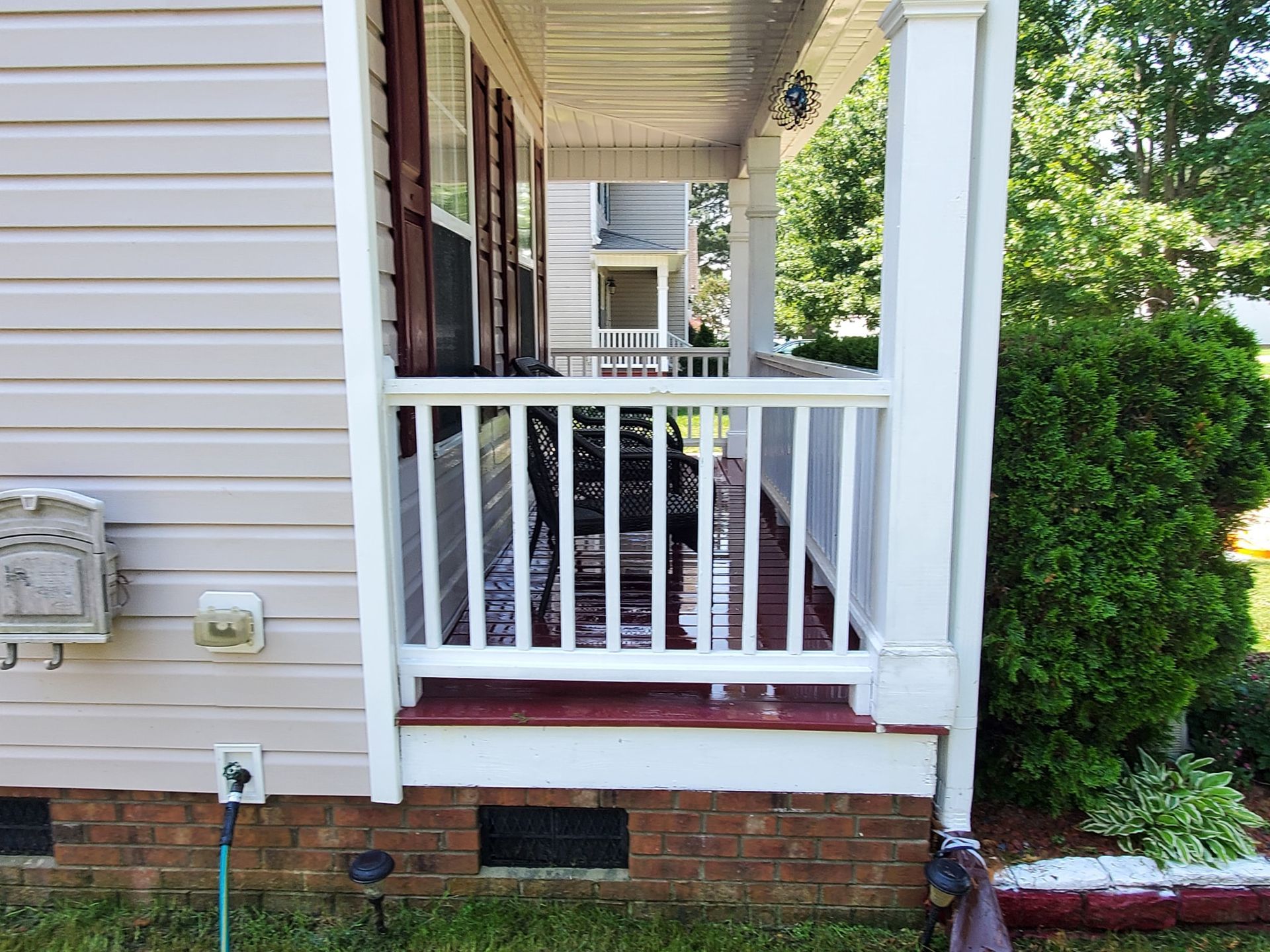 Porch of a house with white railings and columns, red trim, beige siding, and shrubbery.