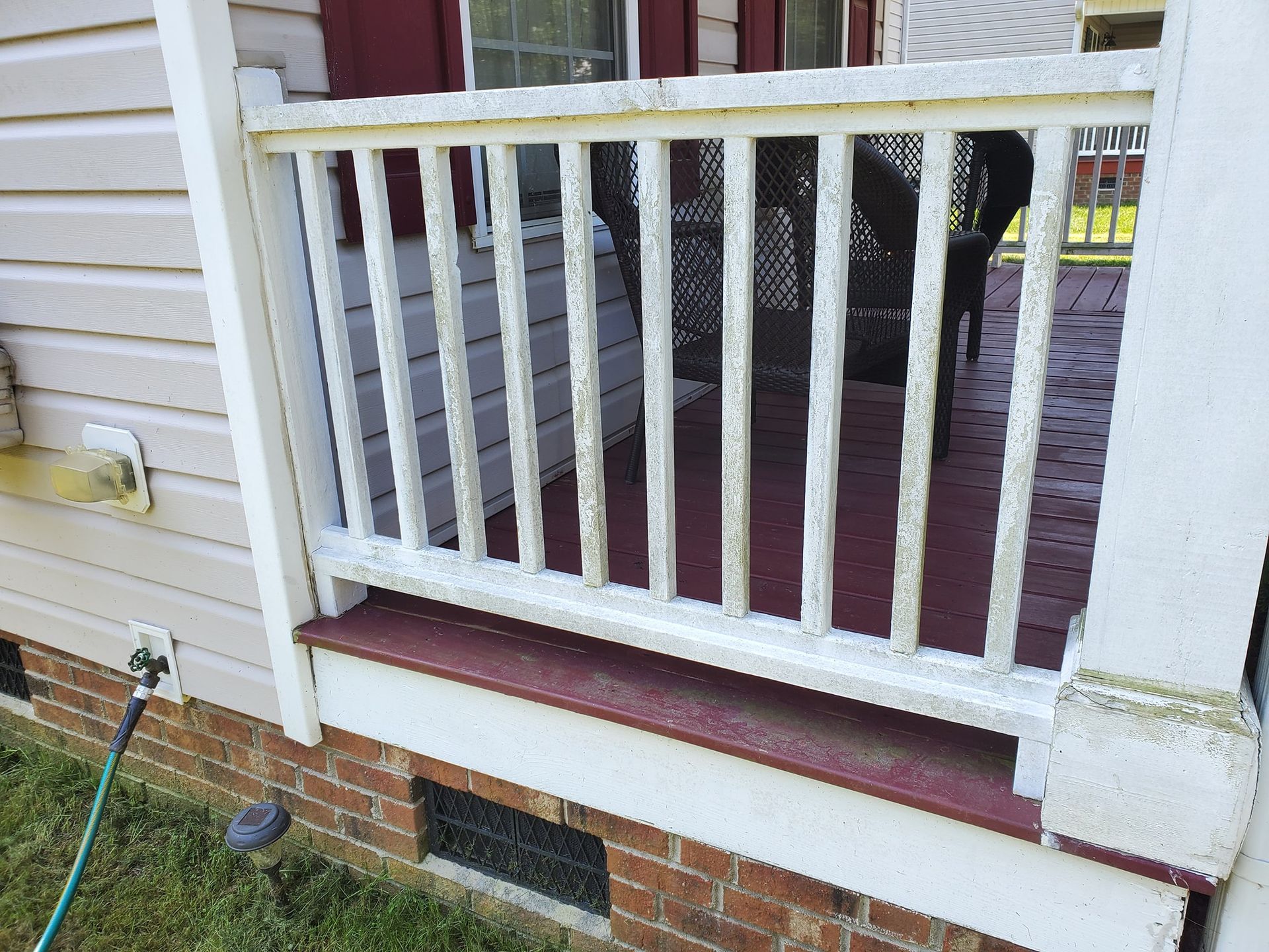 White railing on a maroon deck with a brick base. A hose is on the grass.