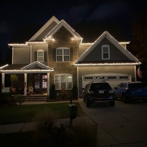 House decorated with white Christmas lights at night, vehicles in the driveway.