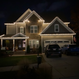 House with holiday lights at night; vehicles parked in driveway.