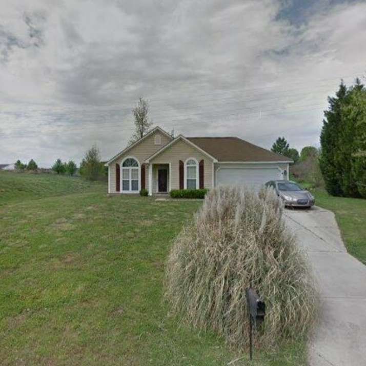 Beige house with brown roof, driveway, car, and overgrown bushes on a grassy lawn under a cloudy sky.