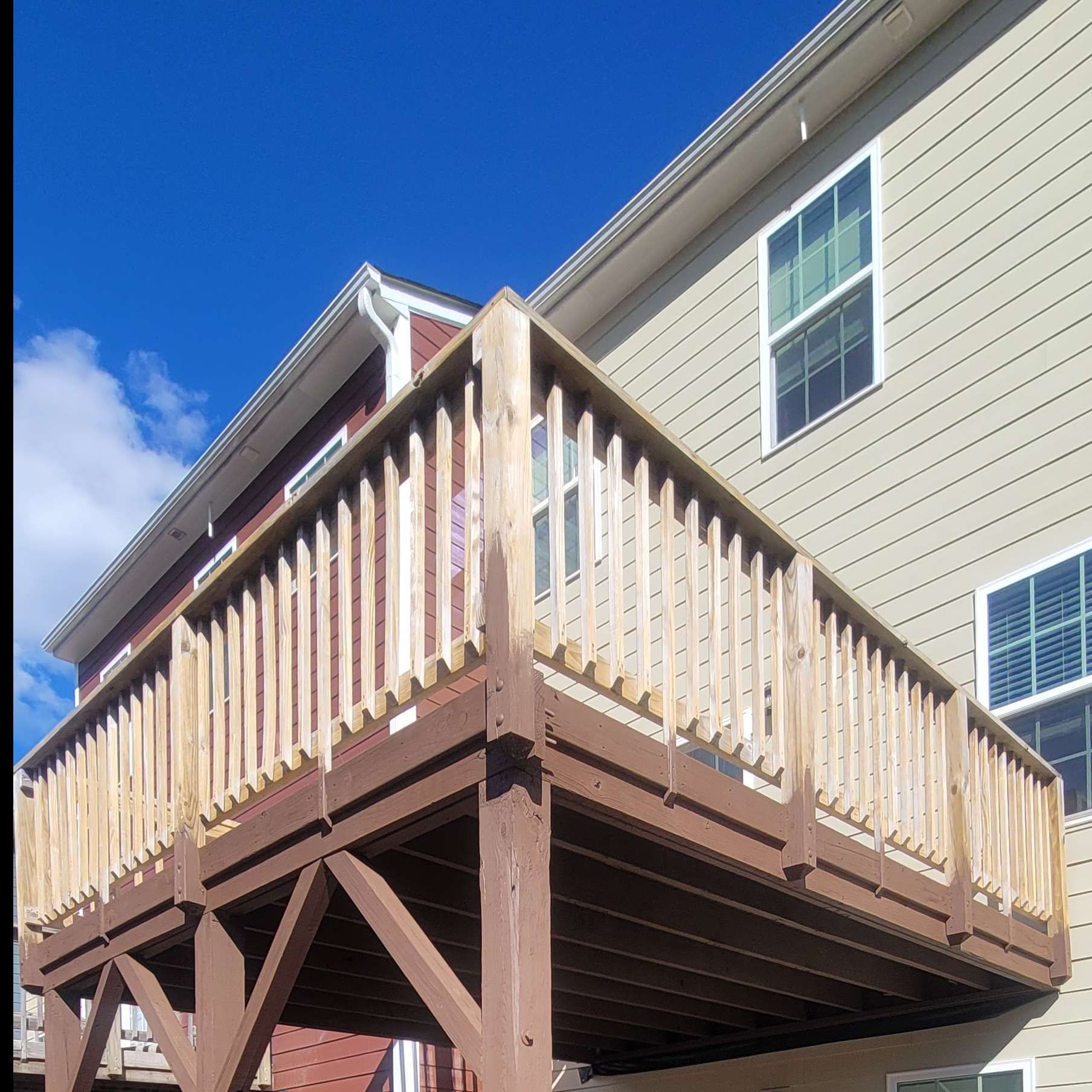 Wooden deck attached to a two-story house with light siding. Sunny day, clear blue sky.