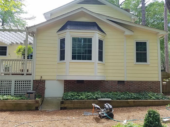 Yellow house with bay window and brick foundation. Lawn equipment sits outside on dirt.