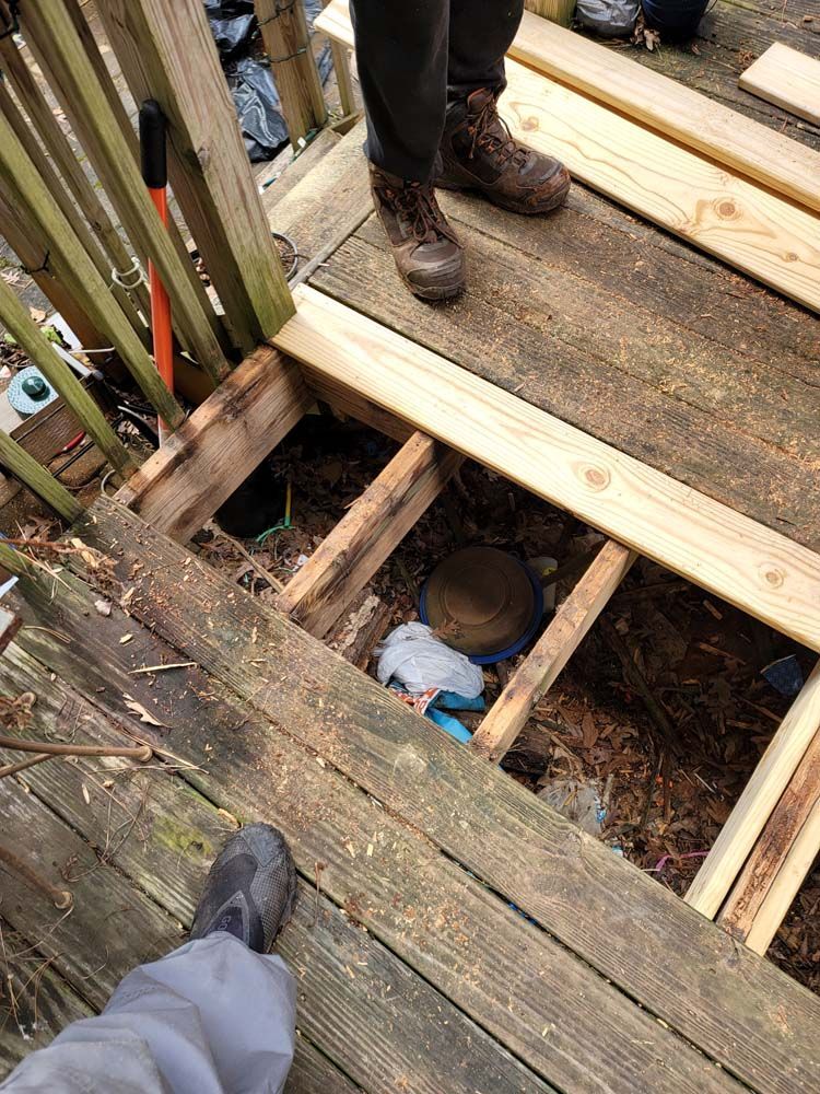 Deck under construction. Person's legs and boots standing on wood boards, revealing deck framing and debris below.