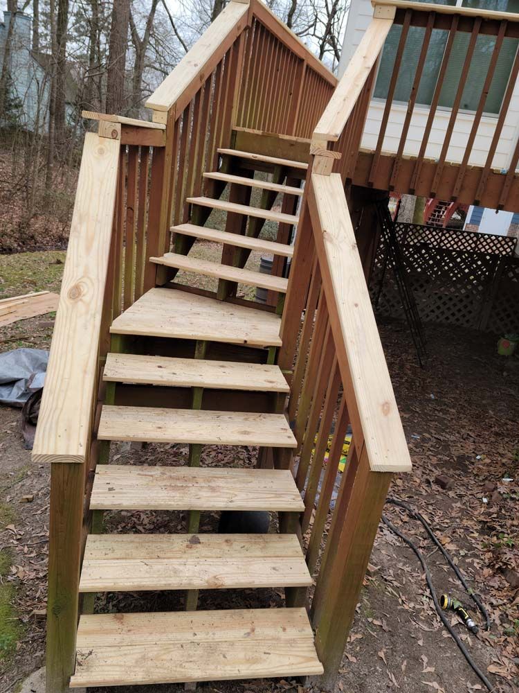 Wooden outdoor staircase leading to a deck. Steps are weathered, surrounded by railings in a backyard setting.
