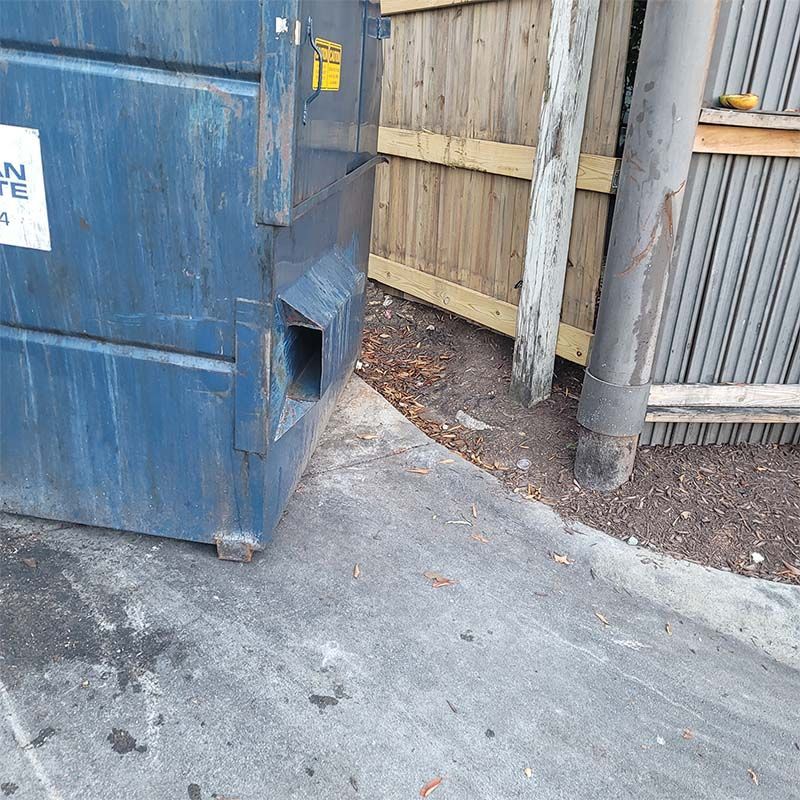 Blue dumpster next to a fence and pole on a concrete surface.