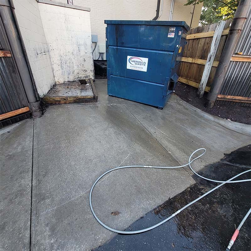 A blue dumpster sits on a wet concrete area, with a hose nearby, suggesting cleaning.