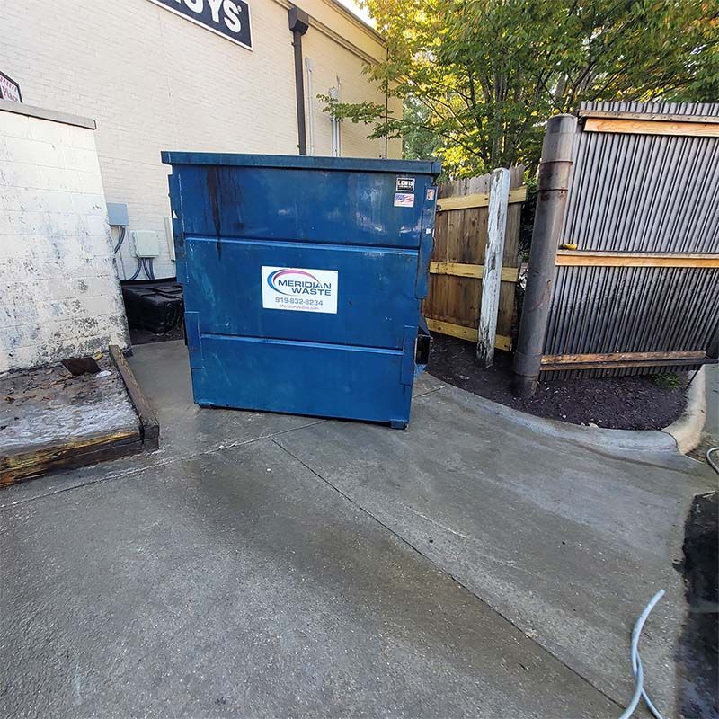 Blue dumpster on a concrete pad, next to a wooden fence and building.