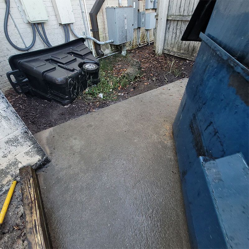 Concrete alley with a tipped black bin, blue dumpster, utility boxes, and white wall.