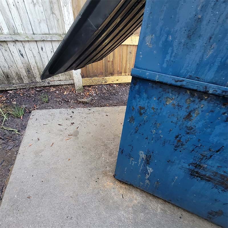 Blue dumpster with open lid on concrete. Wooden fence in background.