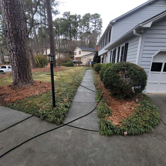 A gray house with a driveway, sidewalk, and shrubs. A black hose is on the sidewalk.