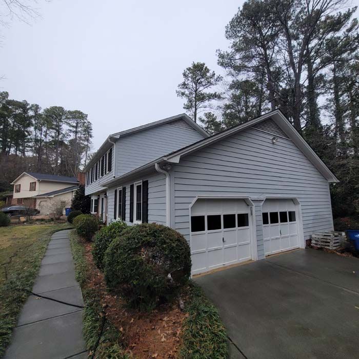 Two-story house with gray siding and garage, black shutters, and green bushes in front, overcast day.