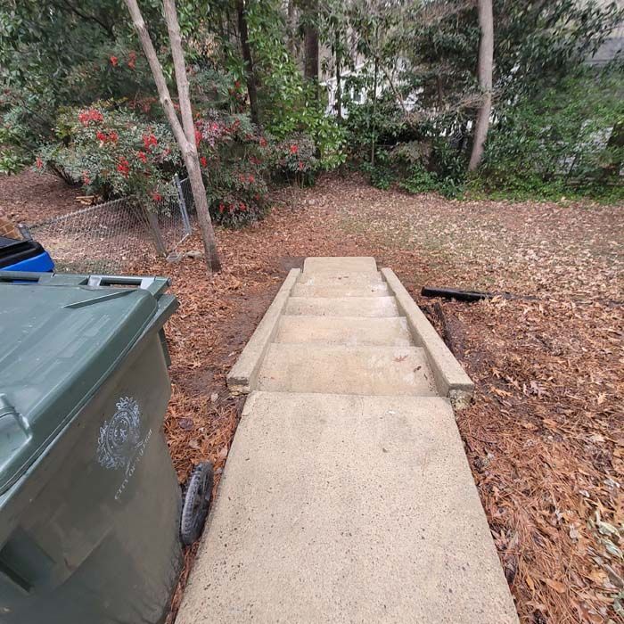 Concrete steps leading up from a walkway, flanked by a trash bin and foliage.