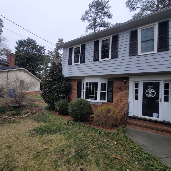 Two-story house with gray siding, brick facade, black shutters, and bay window; yard with shrubs and grass.