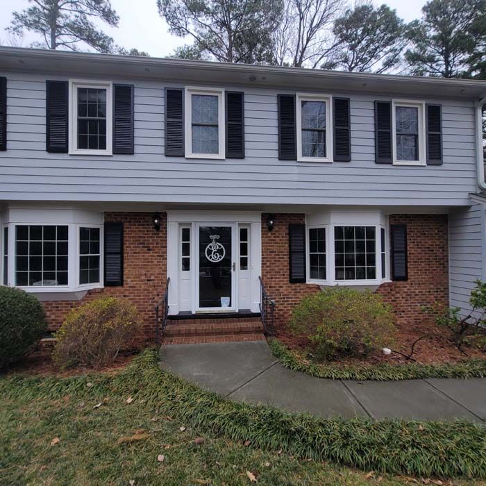 Gray and brick two-story house with black shutters and bay windows; front door with wreath, walkway, and landscaping.