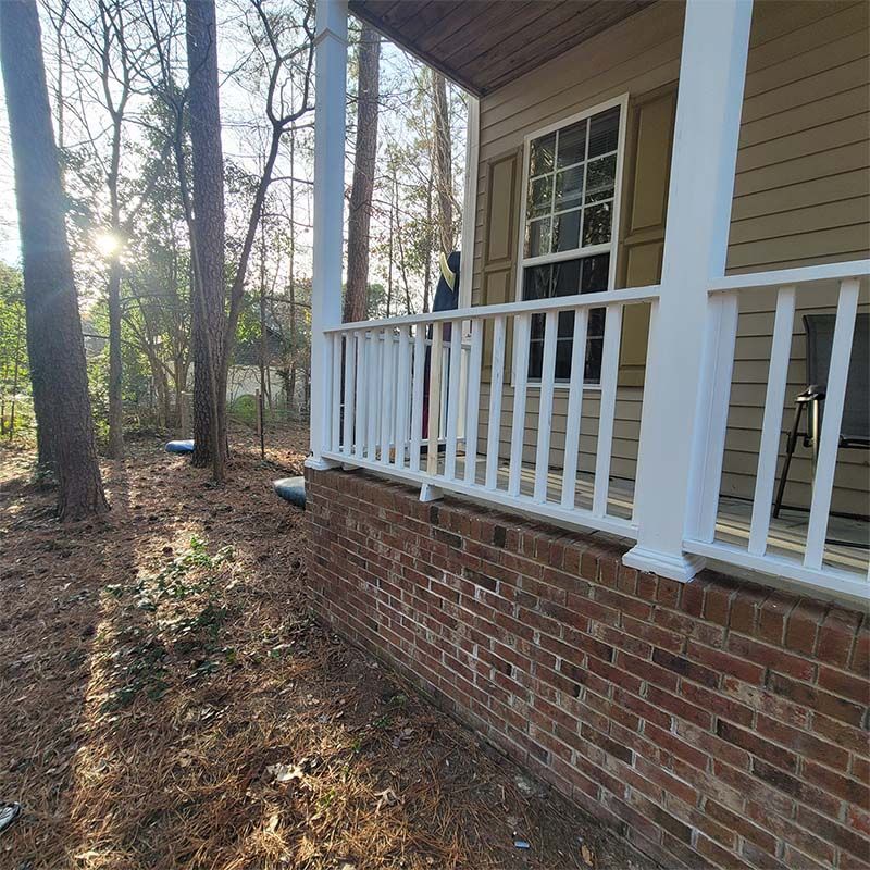 A porch with white railing and columns, brick foundation, surrounded by trees.