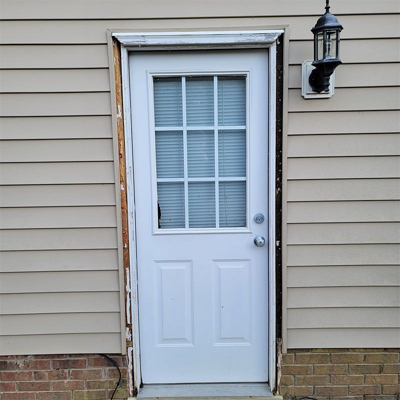 White door with glass panes and warped frame, set into a beige siding exterior.