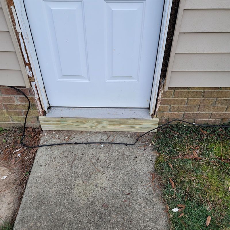 White door with wooden step, concrete path, and grass. A black cable runs across the path.