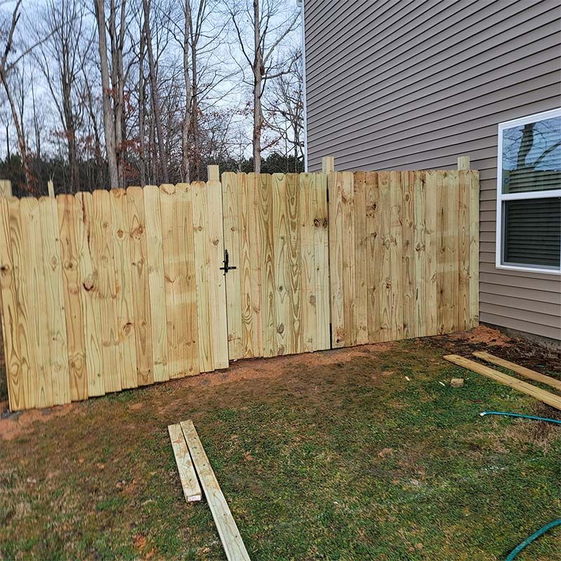Wooden fence with a gate next to a house. Green grass, trees in background.