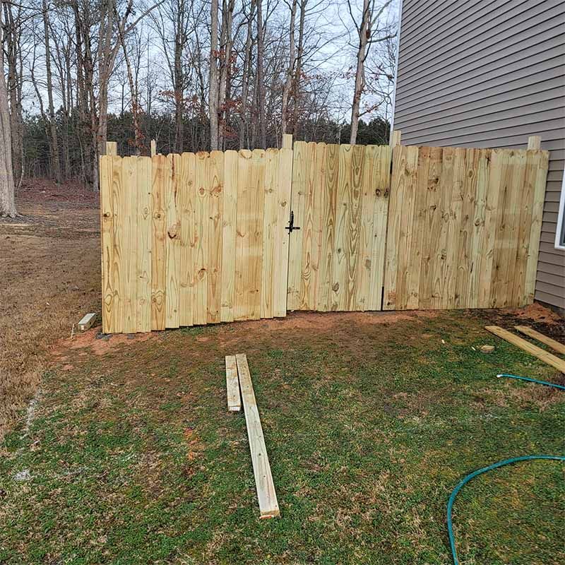 Wooden fence with gate attached to a house. Green grass surrounds. Brown trees in background.