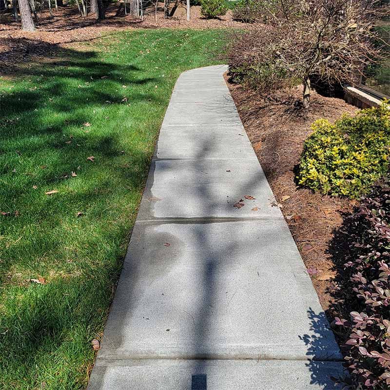 Concrete pathway winding through a grassy lawn and landscaped area. Sunny day with shadows.