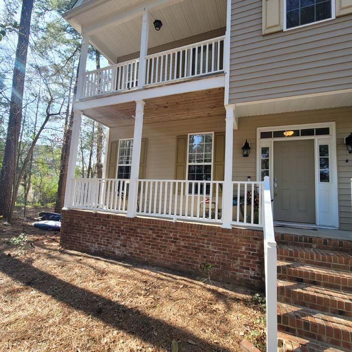 Two-story house with white railings and tan siding. Red brick base. Front porch and a doorway.