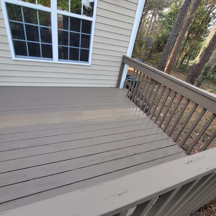A weathered wooden deck painted gray with a railing next to a light tan house with a window.