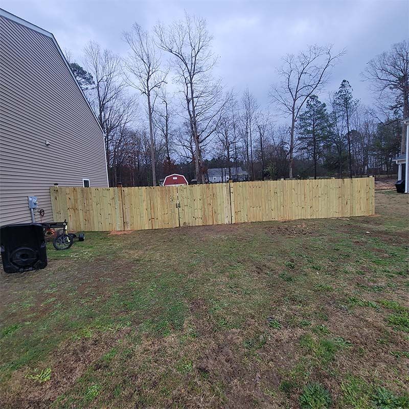 Wooden fence in backyard, surrounding green grassy area. Cloudy sky overhead, trees in the background.