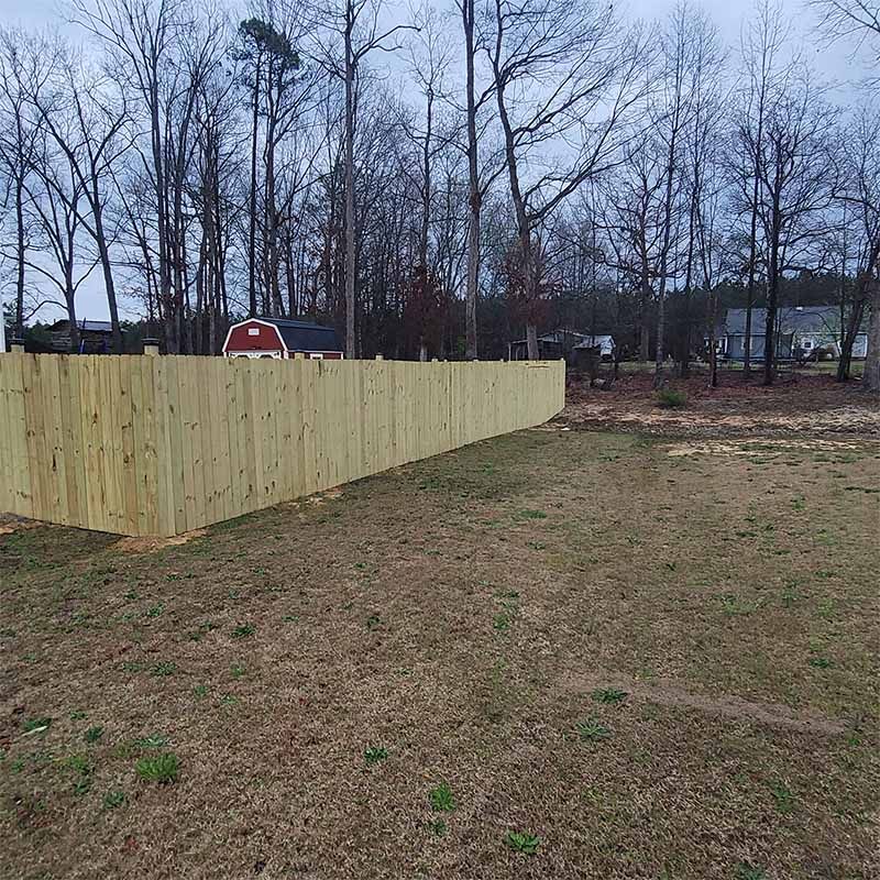 A wooden fence in a backyard with a red building and bare trees in the background.