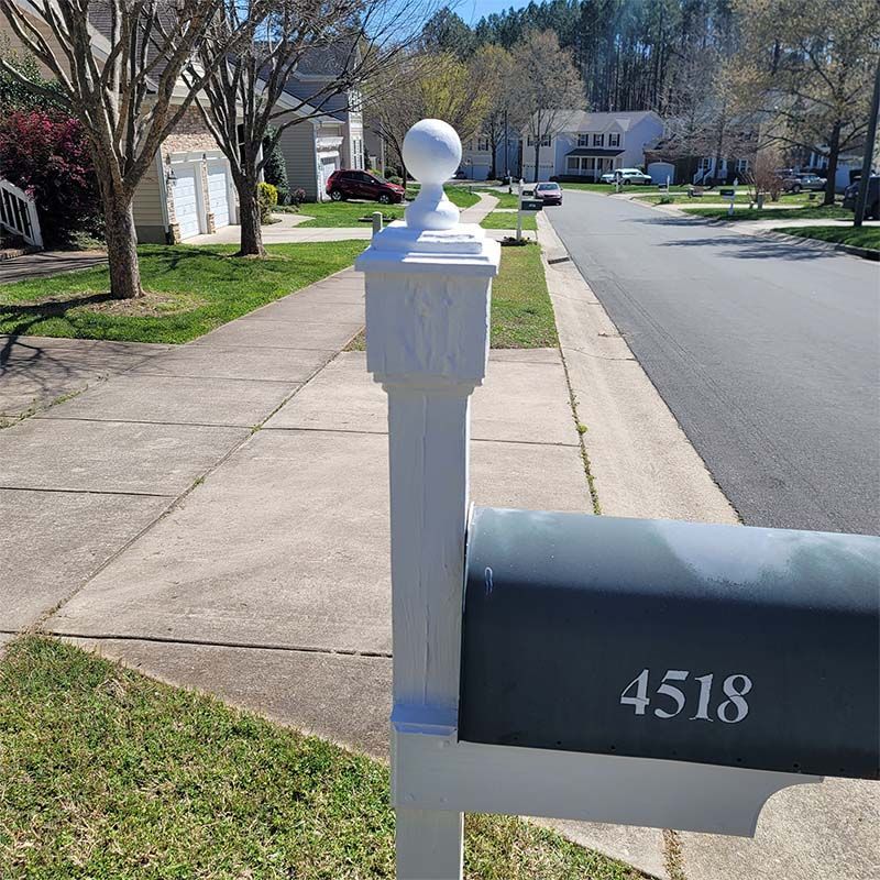 White mailbox post with a decorative top, next to a sidewalk and asphalt road, in a suburban neighborhood.