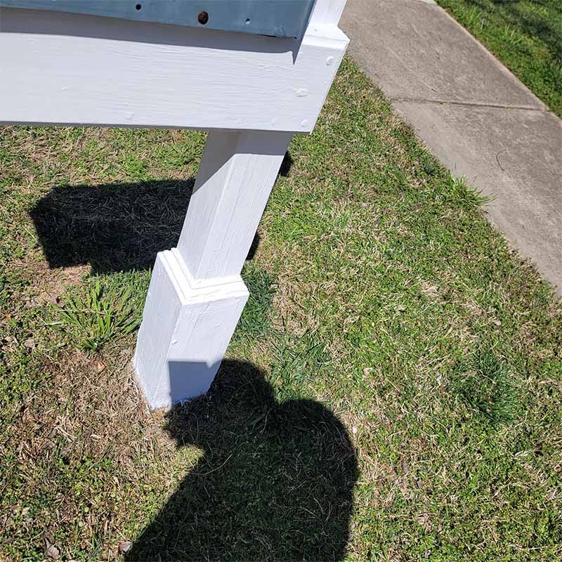 White painted mailbox post on a green lawn next to a sidewalk, casting a shadow.