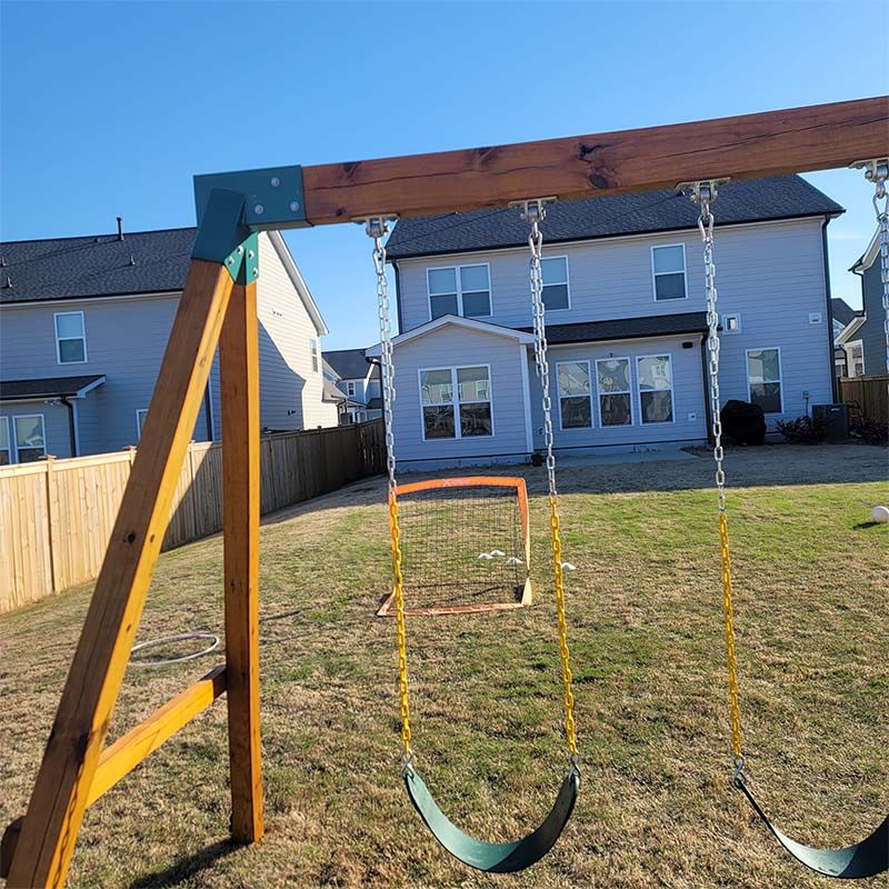 Wooden swing set in a backyard; light blue house and brown fence in background. Sunny day.