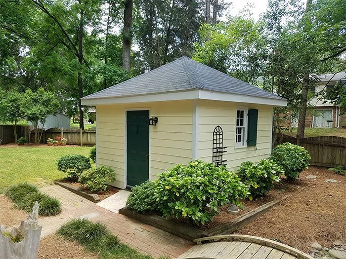 Yellow shed with green door and shutters, surrounded by landscaping and a wooden bridge.