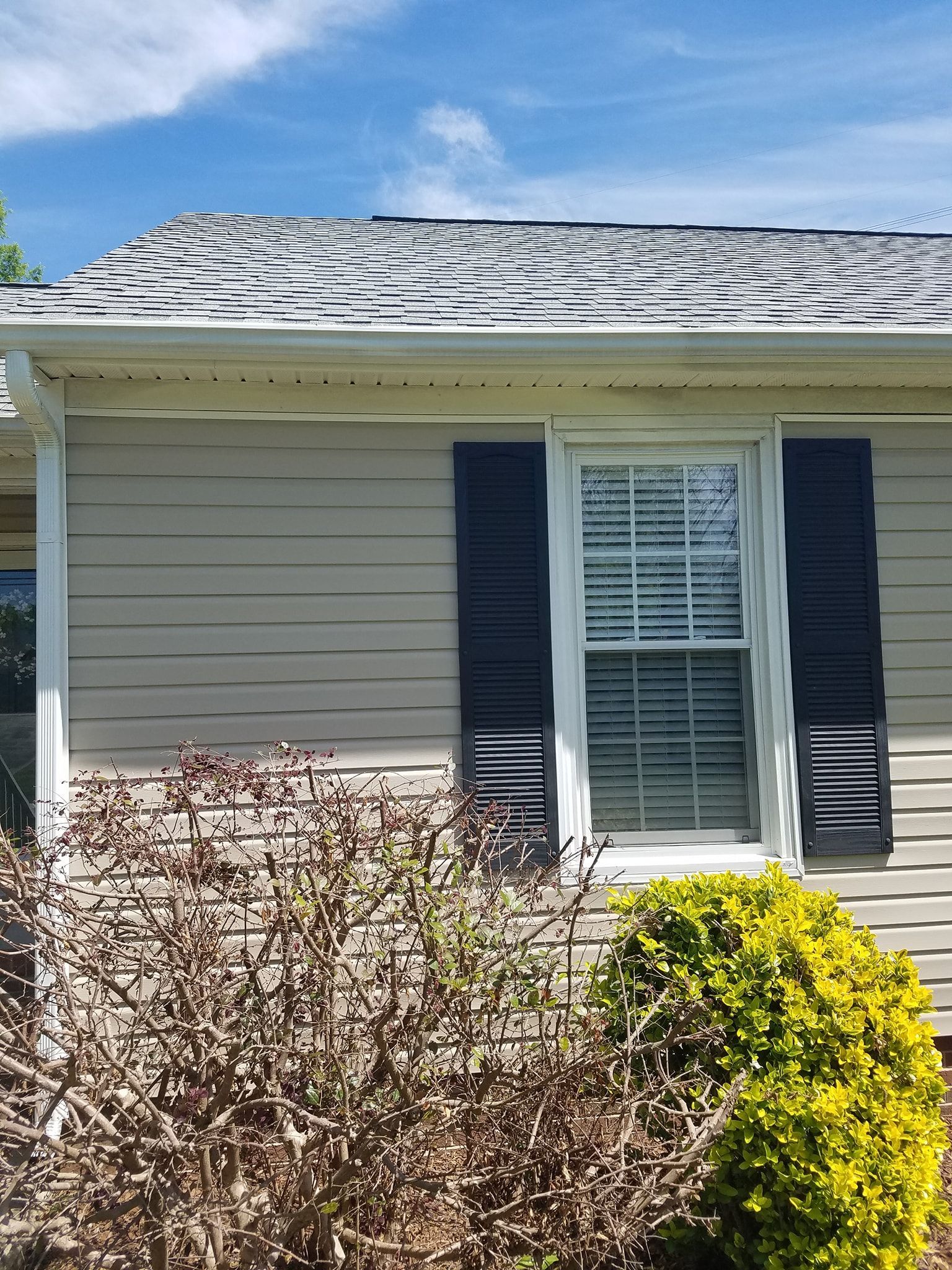 Beige house with a window, dark shutters, and an old, patchy roof.