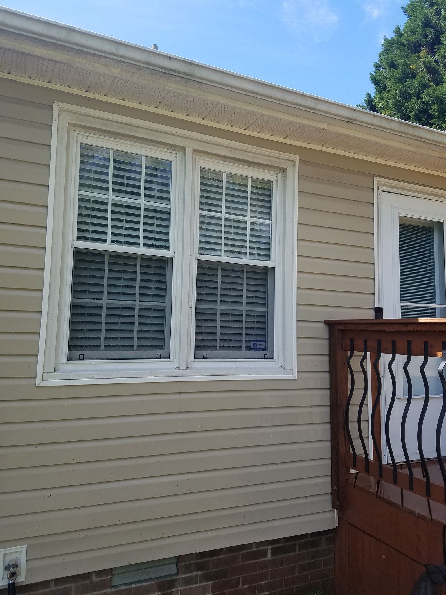 Two white-framed windows with blinds on a beige house, part of a wooden deck visible on the right.